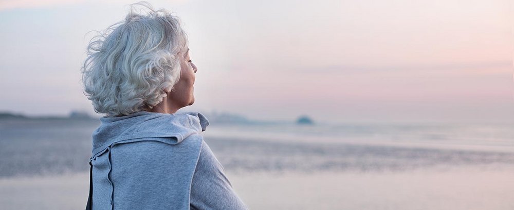 Ältere Dame steht am Strand und blickt aufs Meer