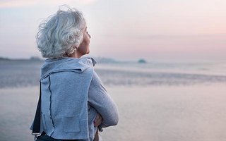 Ältere Dame steht am Strand und blickt aufs Meer